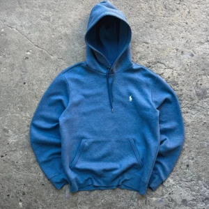 Blå hoodie från Polo Ralph Lauren - Snygg blå hoodie från Polo Ralph Lauren med klassisk logga broderad på bröstet. Tröjan har huva med snörning, känguruficka och ribbade muddar. Materialet är en mjuk blandning av bomull och polyester, perfekt för chill dagar.