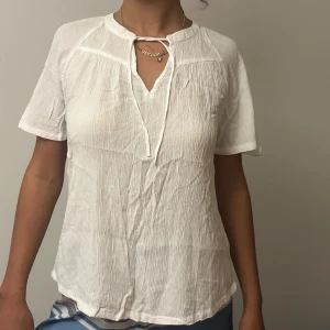 Vit blus med knyt och kort ärm - Luftig vit blus i krinklad bomull med korta ärmar och v-ringning med knytband vid halsen. Blusen har en avslappnad passform och lätt struktur i tyget, perfekt för varma dagar.