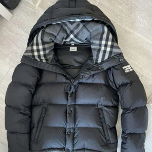 Svart dunjacka från Burberry - Snygg svart dunjacka från Burberry med klassiskt rutigt foder i grå toner. Jackan har huva, dragkedja och knappar framtill samt två fickor med dragkedja. Perfekt för kalla dagar och riktigt stilren look. Storlek XL men passar L