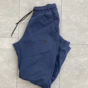 Marinblå Nike tech - Marinblå Nike tech byxor med elastisk midja och snörning. Byxorna har en svart dragkedjeficka på sidan och ribbade muddar vid bensluten. Tillverkade i mjukt bomullsmaterial för maximal komfort. Perfekta för chill eller träning. Finns två små hål, se bilder.