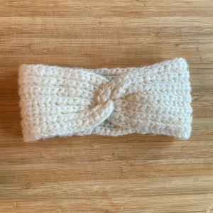 Virkat vitt pannband med twist - Ett chunky virkat pannband i mjuk, vit ull med snygg twist framtill. Perfekt för att hålla öronen varma och samtidigt se stilren ut. Bred modell som sitter skönt på huvudet och ger en mysig vinterlook. Har måtten 20 cm x 9 cm