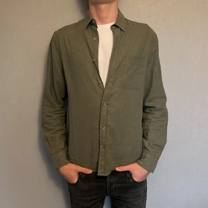 Olivgrön linneskjorta  - Snygg olivgrön skjorta i linne med klassisk krage och knäppning framtill. Perfekt att bära öppen över en t-shirt för en avslappnad look.         (Nypris 500)