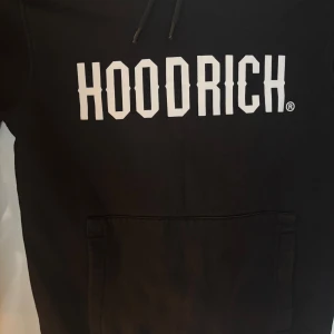 Svart HOODRICH hoodie med tryck - Svart hoodie från HOODRICH med stor vit logga framtill. Klassisk känguruficka, huva med snörning och mjukt material. Perfekt för dig som gillar streetwear och vill ha en stilren look.