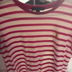 Randig t-shirt från Ralph Lauren - Säljer en snygg t-shirt från Ralph Lauren med vita och röda ränder. Klassisk passform med korta ärmar och rund hals. Ikoniska gula broderade Polo-loggan på bröstet. Perfekt för en clean och stilren look.