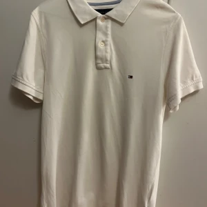 Vit pikétröja Tommy Hilfiger S - Klassisk vit pikétröja från Tommy Hilfiger i slim fit-modell. Tröjan har korta ärmar, krage med två knappar och den ikoniska lilla Hilfiger-loggan broderad på bröstet. Tillverkad i mjuk bomull