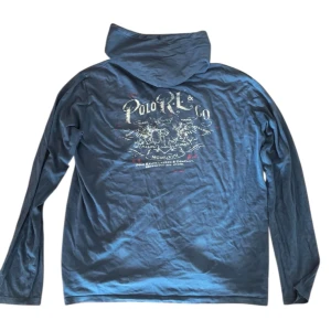 Blå hoodie från Polo Ralph Lauren - Snygg blå hoodie från Polo Ralph Lauren med tryck på ryggen och liten logga på bröstfickan. Tröjan har huva, lång ärm och är tillverkad i mjuk bomull. Perfekt för en avslappnad och cool stil.