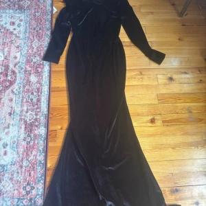 Svart sammetsklänning med lång ärm - Elegant svart långklänning i mjuk sammet med långa ärmar och figurnära passform. Klänningen har en klassisk och stilren look som passar perfekt till speciella tillfällen. Snyggt fall och markerad midja för en sofistikerad siluett.