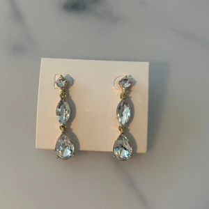 Glittriga örhängen med kristaller - Snygga örhängen med tre stora kristaller i olika former, infattade i guldfärgad metall. Kristallerna är klara och ger ett riktigt glittrigt intryck. Perfekt för att lyfta din look med lite extra bling.