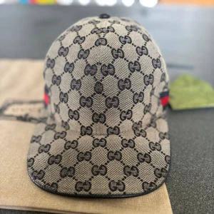 Gucci beige keps med GG-mönster - Snygg beige keps från Gucci med klassiskt GG-monogram i svart över hela kepsen. På sidan finns en ikonisk röd och marinblå rand. Justerbar rem baktill i svart skinn och insida i mörkblått tyg. Perfekt för dig som vill ha en lyxig och trendig look.