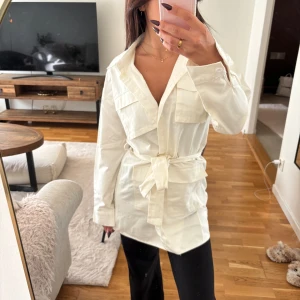 Vit overshirt med bälte och fickor - Säljer en stilren vit overshirt i bomull med knappar framtill, stora bröstfickor och bälte i midjan. Skjortan har lång ärm och klassisk krage, perfekt att styla över ett linne eller t-shirt. Enkel och clean look som passar till många outfits.