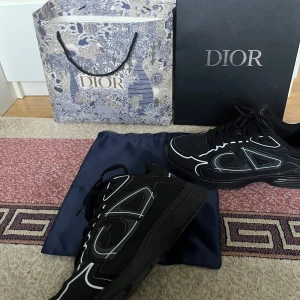 Svarta Dior sneakers med vita detaljer - Säljer ett par svarta Dior sneakers med vita kontrastdetaljer och tydlig CD-logga på sidan. Skorna har snörning och en chunky sula för en modern look. Kommer med originalpåse, kartong och Dior-påse. Perfekta för dig som vill sticka ut med lyxig streetstyle.