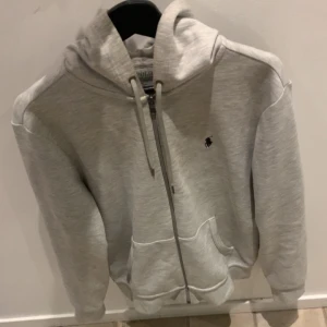 Ralph Lauren zip hoodie  - Snygg ljusgrå hoodie från Polo Ralph Lauren med dragkedja framtill, huva med snörning och klassisk broderad logga på bröstet. Mjuk bomullsblandning och två fickor framtill. Perfekt för chill dagar eller när du vill ha en clean look.