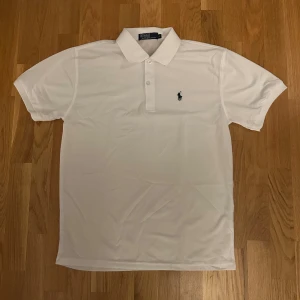 Vit pikétröja från Ralph Lauren - Klassisk vit pikétröja från Ralph Lauren med korta ärmar och broderad logga på bröstet. Tröjan har krage, två knappar framtill och är tillverkad i bomull. Perfekt för en clean och stilren look.