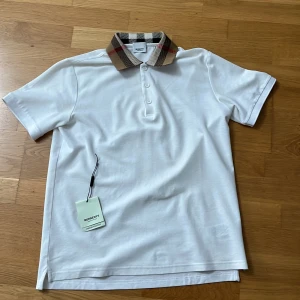 Vit Burberry piké med rutig krage - Vit pikétröja från Burberry med klassisk rutig krage i beige, svart och rött. Är använd 1-3 gånger. Lite stor i storlek