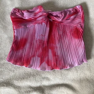 Plisserad rosa-röd tubtopp från SHEIN - Säljer en snygg plisserad tubtopp från SHEIN i storlek S. Toppen är axelbandslös och har en mix av rosa och rött i ett unikt mönster. Materialet är lätt och glansigt, perfekt för att sticka ut. Toppen har twistad detalj framtill och är croppad i modellen.