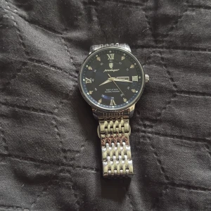 Silver klocka  - Stilren klocka från Pedagar med svart urtavla och romerska siffror i guld. Armbandet är i blankt, flätat rostfritt stål och boetten är rund. Urtavlan har små kristalldetaljer vid varje timmarkering och datumvisning. Perfekt för dig som gillar klassisk design med en modern touch.