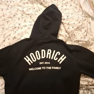 Svart zip-hoodie från Hoodrich - Svart hoodie från Hoodrich med vit text både fram och bak. Tröjan har huva med snörning, dragkedja hela vägen och kängurufickor. Snygg streetstil med trycket 'Welcome to the family' och 'EST. 2014'. Perfekt för dig som gillar urban mode.