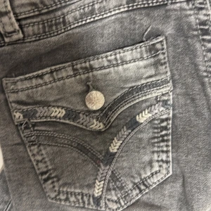 Grå lowrise bootcut jeans med broderad ficka - Snygga grå jeans med bootcut passform och coola broderade detaljer på bakfickan. Jeansen har en unik knapp och kontrastsömmar som ger extra edge. Perfekta för dig som vill sticka ut lite extra. Materialet är klassisk denim.