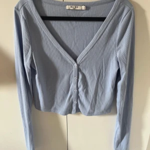 Ljusblå ribbad topp från NA-KD - Säljer en ljusblå ribbad topp från NA-KD med v-ringning och knappar framtill. Toppen har lång ärm och croppad passform