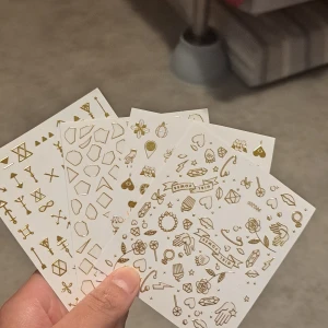 Guldiga nagelstickers i olika motiv - Fyra ark med nagelstickers i metallic guld, fyllda med olika motiv som hjärtan, blommor, geometriska former, pilar och smycken. Perfekta för att skapa unika och kreativa nageldesigner. Självhäftande och enkla att använda på både naturliga och målade naglar.