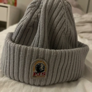 Grå ribbstickad mössa från Parajumpers - Snygg grå ribbstickad mössa från Parajumpers med uppvikt kant och klassisk PJS-logga framtill. Perfekt för kalla dagar och enkel att matcha med olika outfits. Tillverkad i mjukt material som håller dig varm och bekväm.