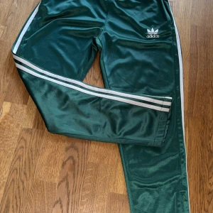 Gröna Adidas träningsbyxor med ränder - Mörkgrönaw träningsbyxor från Adidas med klassiska vita ränder längs sidorna och Adidas-logga på låret. Byxorna har en glansig finish och är tillverkade i syntetmaterial, perfekta för en sportig look.