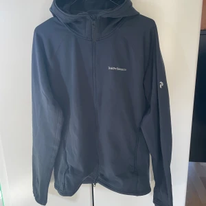 Marinblå  midlayer zip Peak Performance XL - Marinblå hoodie från Peak Performance i storlek XL. Tröjan har dragkedja framtill, huva och två sidofickor med dragkedja. Perfekt för kyliga dagar och har en stilren logga på bröstet samt ärmen. Materialet är mjukt och känns sportigt.