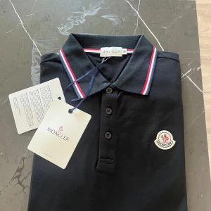 Svart Moncler piké med logga - Svart Moncler pikétröja i bomull med klassisk krage och knäppning framtill. Kragen och ärmsluten har snygga ränder i rött och vitt. Ikonisk Moncler-logga på bröstet. Perfekt för dig som vill ha en stilren och sportig look.