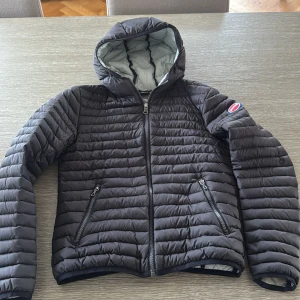 Svart pufferjacka från Colmar - Snygg svart pufferjacka från Colmar med huva och två dragkedjeförsedda fickor. Jackan har ett quiltat mönster och klassisk Colmar-logga på ärmen. Barnstorlek 12Y 154cm. 