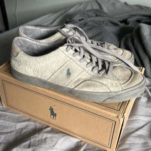 Grå mockasneakers från Polo Ralph Lauren - Snygga sneakers i beige mocka från Polo Ralph Lauren med klassisk logga på sidan. Skorna har grå snörning, platt sula och rund tå. Perfekta för dig som gillar stilrena och tidlösa sneakers med en touch av premiumkänsla.