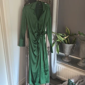 Grön omlottklänning Gina Tricot - Snygg lång grön omlottklänning från Gina Tricot i glansigt material. Klänningen har långa ärmar, breda manschetter, krage och knyt i midjan för en smickrande siluett. Perfekt för dig som vill sticka ut med en elegant look.
