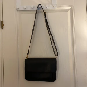 Svart axelväska i skinn med guldiga detaljer - Det är den perfekta väskan inför hösten! Den har en perfekt passform och passar perfekt in till höstens trender då den är svart med guld detaljer och det får plats minst en bok i den.  Har även ett minde fack inuti för plånbok. Ett litet fack på utsidan också som kan kan lägga mobilen i till exempel.