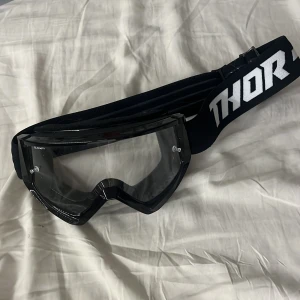 Svarta Combat Youth goggles från Thor - Säljer ett par svarta Combat Youth goggles från Thor med genomskinligt visir och bred elastisk rem med vit Thor-logga. Perfekta för motocross eller liknande aktiviteter. Ramen är i plast och insidan har mjuk skumvaddering för extra komfort.