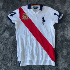 Ralph Lauren Polo - Vit t-shirt från Polo Ralph Lauren med korta ärmar, stor mörkblå polospelare på bröstet, röd diagonal rand och broderad patch. Mörkblå krage med röd/vit kant, stora siffror 14 på rygg och ärmar. Materialet är bomull, perfekt för en sportig look. ”Chief Keef style”