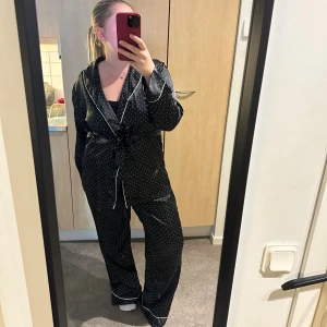 Svarta prickiga pyjamasbyxor o tröja i satin - Snyggt pyjamasset från Lindex i storlek L  i glansigt satinmaterial med vita små prickar. Byxorna har en avslappnad passform och resår i midjan för extra komfort. Och tröjan har ett band i midjan. Jättesköna o ha hemma! Använt fåtal gånger. Säljer för 80kr.