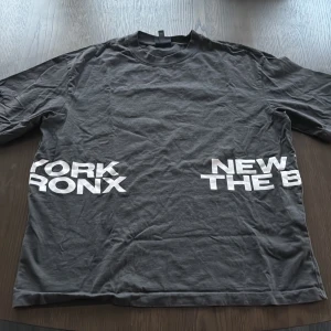 Svart loose fit t-shirt från H&M - Svart t-shirt från H&M i loose fit med vit texttryck på både fram- och baksidan där det står 'NEW YORK BRONX THE BX'. Klassisk rund hals och korta ärmar. Skön och avslappnad stil, perfekt till jeans eller shorts.