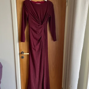 Vinröd glittrig långklänning - Säljer en vinröd långklänning med glittrigt tyg och lång ärm. Klänningen har en snygg drapering framtill och rund halsringning. Perfekt för dig som vill sticka ut med en elegant och trendig look.