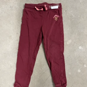Vinröda Hogwarts mjukisbyxor - Mysiga vinröda mjukisbyxor med Hogwarts-logga i guld och rosa på ena benet. Byxorna har ribbad midja med rosa snörning och muddar vid bensluten. Perfekta för chill eller Harry Potter-fans som vill ha något bekvämt.