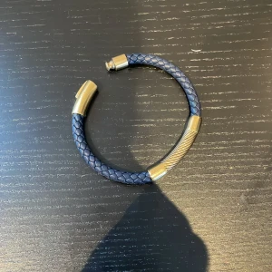 Blått flätat armband med gulddetaljer - Snyggt armband i blått flätat material med eleganta guldfärgade metalldetaljer. Perfekt accessoar för att lyfta din outfit med en stilren touch. Magnetlås för enkel användning.