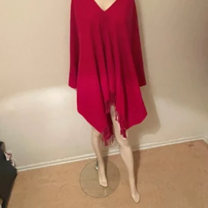 Mörk rosa ponchoklänning med fransar - En mörk rosa ponchoklänning med v-ringning och långa ärmar. Klänningen har en lös passform och är dekorerad med fransar längs nederkanten. Perfekt för dig som gillar en avslappnad och trendig stil.