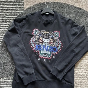 Svart sweatshirt från Kenzo med tiger - Svart sweatshirt från Kenzo med ikonisk broderad tiger i flera färger och texten 'Kenzo Paris' framtill. Tröjan har rund hals, ribbade muddar och är långärmad. Perfekt för dig som gillar statement-plagg och coola detaljer.