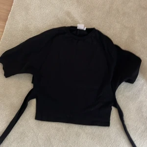 Svart knyt toppfrån Nelly XS - Svart omlottopp från Nelly i storlek XS. Toppen har korta ärmar och knytband i midjan som ger en snygg siluett. Enkel och stilren design som passar perfekt till jeans eller kjol. Mjukt material som känns skönt mot huden.