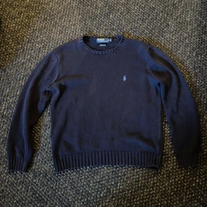 Mörkblå tröja från Polo Ralph Lauren - Klassisk mörkblå tröja från Polo Ralph Lauren i 100% bomull. Tröjan har rund halsringning, ribbade muddar och det ikoniska broderade logotypen på bröstet. Perfekt för dig som gillar stilren och tidlös design.