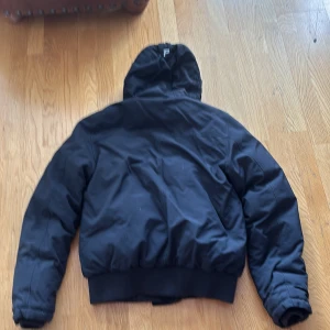 Svart dunjacka Peak Performance L väldigt liten i storleken så skulle säga SS - Svart dunjacka från Peak Performance med huva och bred mudd nertill. Jackan har två stora fickor framtill med lock och dragkedja, samt diskret logga på ena fickan. Perfekt för kalla dagar och har en klassisk, sportig look.