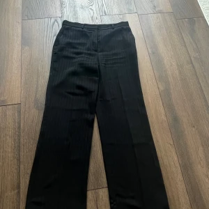 Svarta kostymbyxor med smala ränder - Svarta kostymbyxor med tunna vita ränder. Klassisk rak passform, bälteshällor och fickor fram samt en bakficka med knapp. Storlek 40.