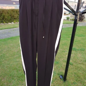 Svarta mjukisbyxor med vita ränder Amisu - Svarta mjukisbyxor från Amisu i storlek 36, med vita ränder längs sidorna och snörning i midjan. Byxorna har en loose passform och är perfekta för chill dagar. Materialet känns mjukt och skönt mot huden. Snygg sportig look med raka ben och diskreta fickor.