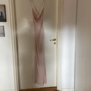 Ljusrosa långklänning från ASOS Design - Supersnygg ljusrosa långklänning från ASOS Design med tunna axelband och draperad urringning. Klänningen har öppen rygg med korsade band och är gjord i ett lätt, flytande material som ger en elegant siluett.