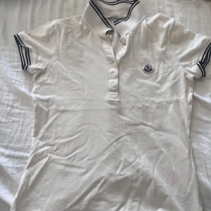 Vit Moncler pikétröja - Vit pikétröja från Moncler med klassisk krage och blå ränder på krage och ärmslut. Tröjan har korta ärmar, knappslå med fyra knappar och Moncler-logga broderad på bröstet. Tillverkad i bomull för en skön och stilren look.