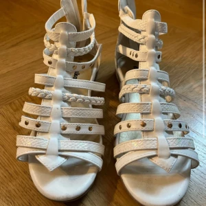 Vita sandaler från Din Sko, strl 36 - Snygga vita sandaler med flera remmar, flätade detaljer och små nitar. Dragkedja bak för enkel påtagning. Platt sula och öppen tå, perfekt för varma dagar. Tillverkade i syntetmaterial med cool design som sticker ut.