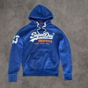 Blå Superdry hoodie med tryck - Blå hoodie från Superdry med vit och orange logga framtill och stort tryck på ena ärmen. Tröjan har huva med vita snören och en stor magficka. Mjuk insida och klassisk passform, perfekt för chill dagar.